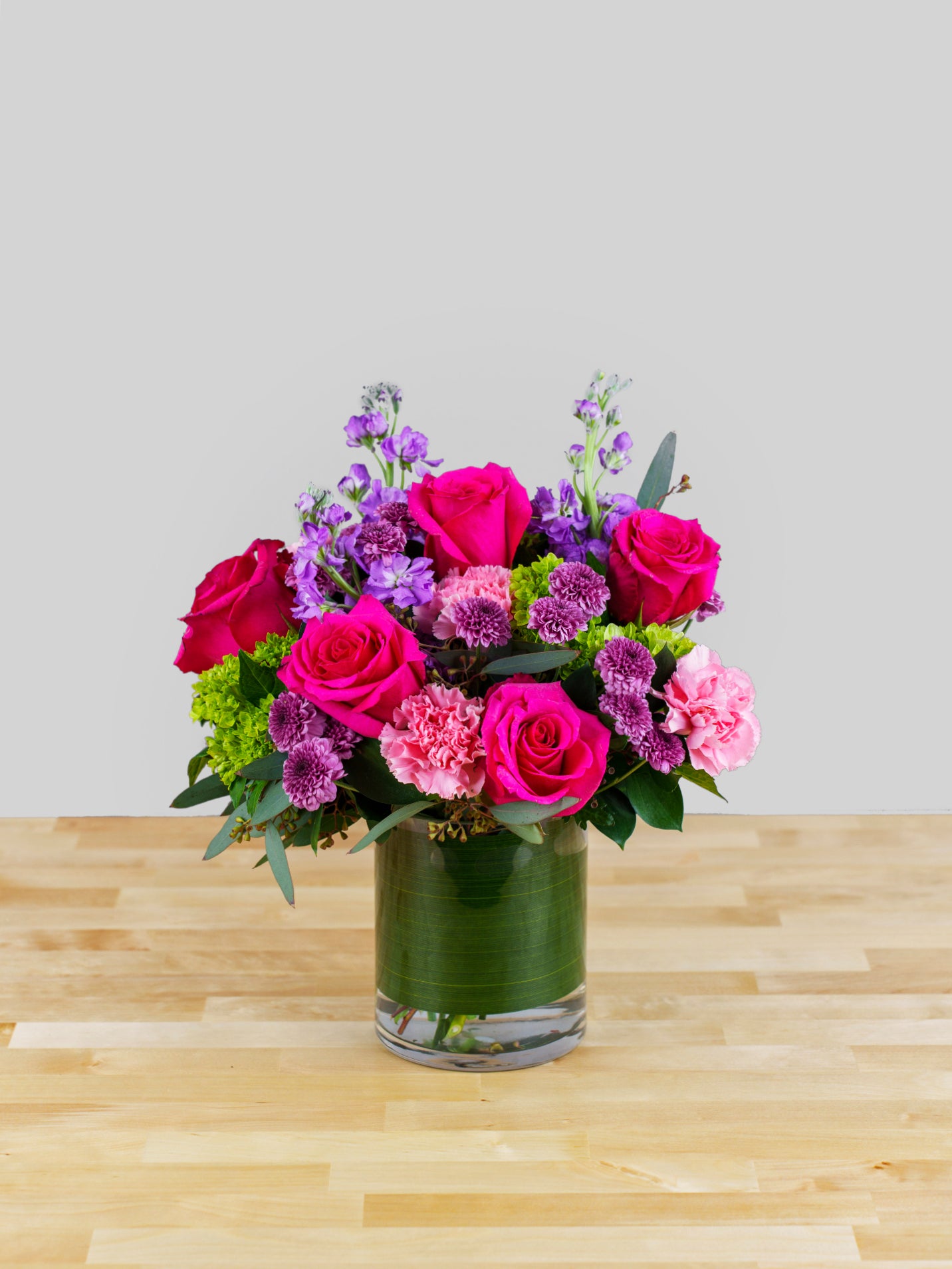 A floral arrangement featuring hot pink roses, lavender buttons, pink carnations, and green hydrangea in a clear vase, placed on a wooden surface.