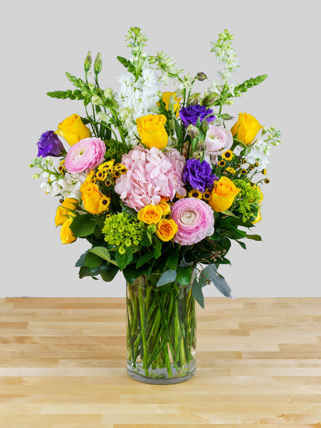 A floral arrangement featuring a variety of flowers including white, yellow, and pink, with green foliage, presented in a clear vase.